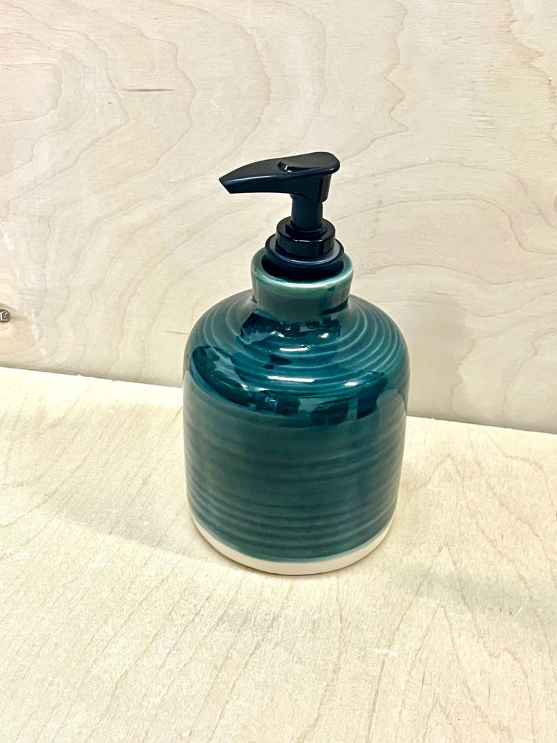 Pu&ograve; includere: Un dispenser di sapone cilindrico in ceramica con una smaltatura color verde acqua profondo e texture a coste orizzontali. La base &egrave; in ceramica bianca non smaltata e la parte superiore &egrave; dotata di un meccanismo a pompa in plastica nera.