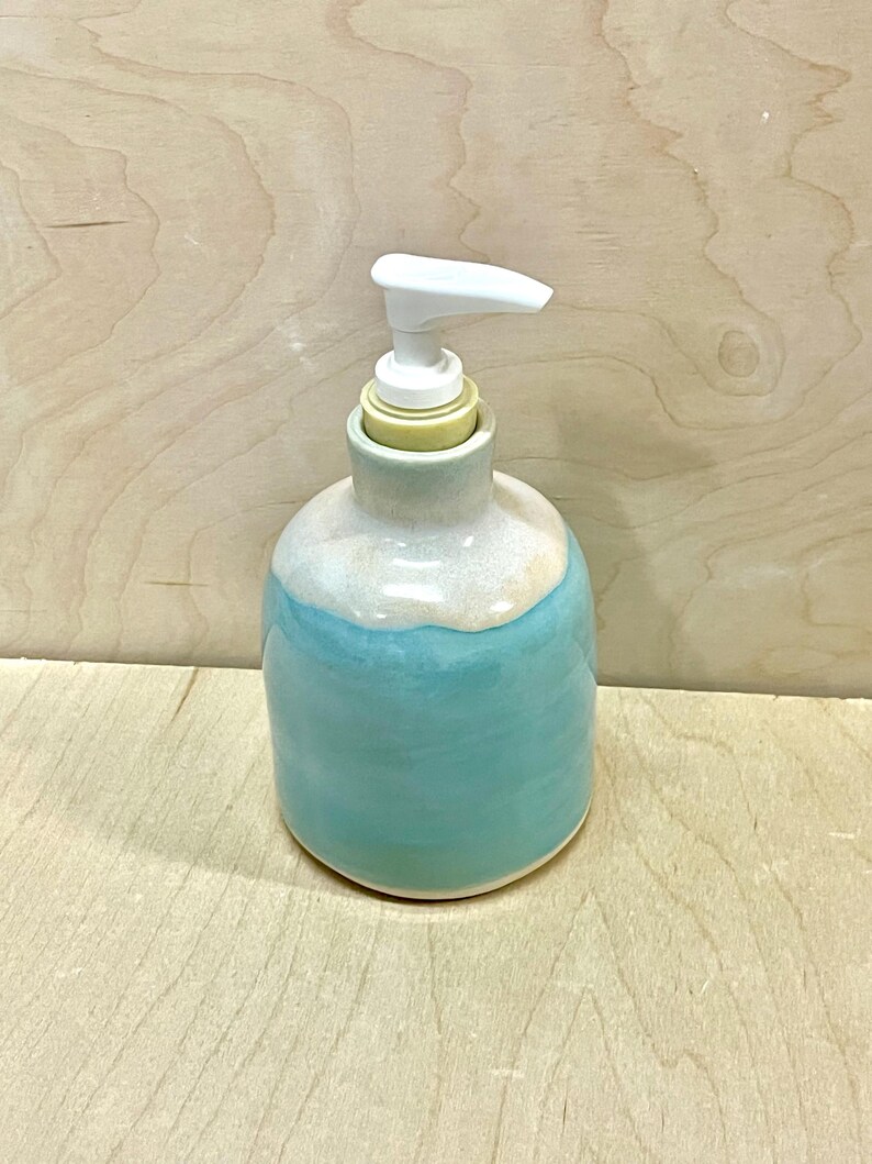Pu&ograve; includere: Un dispenser per sapone in ceramica con corpo arrotondato, caratterizzato da una smaltatura turchese lucida nella parte inferiore e una smaltatura bianca liscia sul collo superiore. Il dispenser &egrave; dotato di una pompa in plastica bianca.