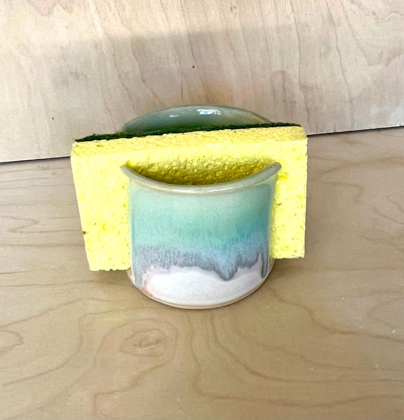 May include: A ceramic sponge holder with a cylindrical shape and a smooth glaze featuring a gradient of light green, teal, and white. A rectangular yellow sponge with a dark green scouring pad is inserted into the holder's center slot.