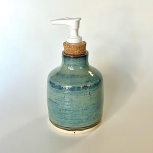Pu&ograve; includere: Un dispenser per sapone in ceramica con una smaltatura lucida blu e verde caratterizzata da linee di texture orizzontali. Il contenitore ha un corpo arrotondato, un tappo in sughero sul collo e una pompa in plastica bianca sulla sommit&agrave;.