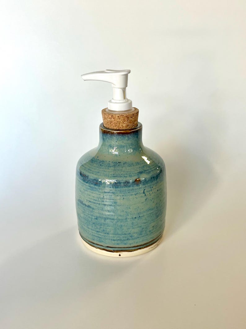 Pu&ograve; includere: Un dispenser per sapone in ceramica con una smaltatura lucida blu e verde caratterizzata da linee di texture orizzontali. Il contenitore ha un corpo arrotondato, un tappo in sughero sul collo e una pompa in plastica bianca sulla sommit&agrave;.