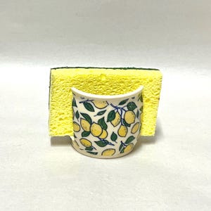 May include: A ceramic sponge holder with a white base and a repeating pattern of yellow lemons and green leaves. A rectangular yellow kitchen sponge with a dark green scouring layer is inserted into the holder, standing upright.