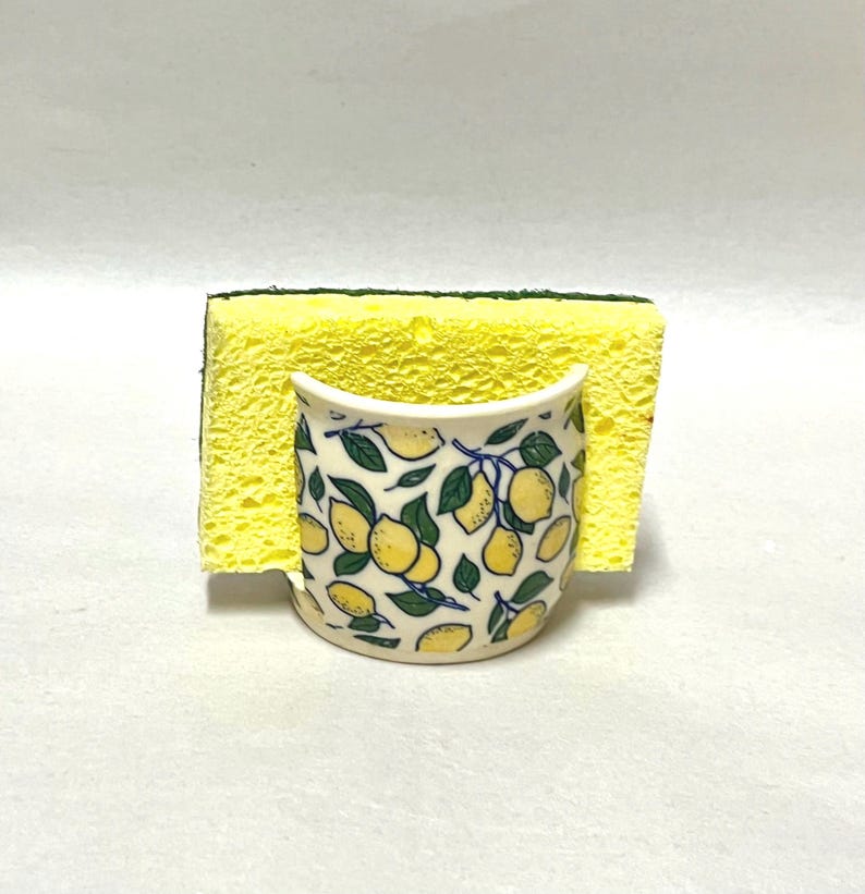 May include: A ceramic sponge holder with a white base and a repeating pattern of yellow lemons and green leaves. A rectangular yellow kitchen sponge with a dark green scouring layer is inserted into the holder, standing upright.