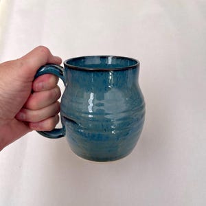 May include: A handmade ceramic mug with a blue glaze. The mug has a rounded body, a sturdy handle, and a dark rim. The mug is held by a hand against a white background. The mug is approximately 4 inches tall.