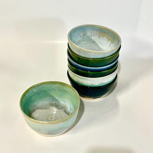 May include: A set of ceramic bowls featuring a reactive glaze in shades of teal, green, and blue. One bowl sits separately in the foreground, while four additional bowls are stacked vertically behind it, showcasing smooth, rounded rims and glossy finishes.