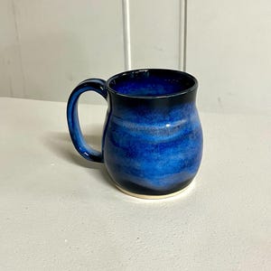May include: A handmade ceramic mug with a deep blue glaze. The mug features a rounded body, a curved handle, and a black rim. The base of the mug is a light cream color. The mug is designed for hot beverages.