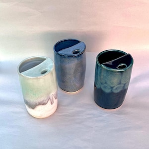 May include: Three cylindrical ceramic toothbrush holders with a semi-circular top shelf featuring a circular cutout. The left holder is light green and white, the centre is medium blue, and the right holder is a deep, dark teal blue.