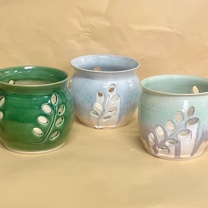 May include: Three ceramic pots in shades of green, blue, and purple. Each pot has a cylindrical shape with a series of leaf-shaped cutouts. The pots are glazed and have a glossy finish, perfect for home decor.