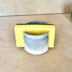 May include: A ceramic sponge holder with a smooth, light blue and white glazed finish. The holder features a curved, semi-circular front design that securely supports a rectangular yellow sponge with a dark green scouring pad layer on top.