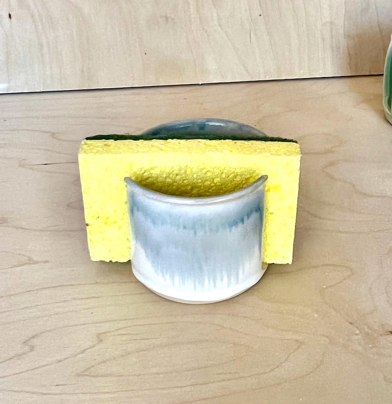 May include: A ceramic sponge holder with a smooth, light blue and white glazed finish. The holder features a curved, semi-circular front design that securely supports a rectangular yellow sponge with a dark green scouring pad layer on top.