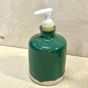Pu&ograve; includere: Un dispenser per sapone cilindrico in ceramica con una smaltatura verde scuro lucida sulla parte superiore e una base opaca color bianco sporco non smaltata. Il dispenser presenta un meccanismo a pompa in plastica bianca con un beccuccio piatto.