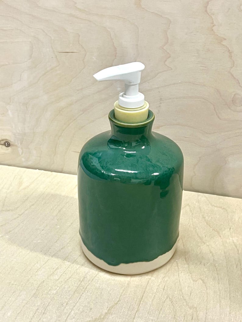 Pu&ograve; includere: Un dispenser per sapone cilindrico in ceramica con una smaltatura verde scuro lucida sulla parte superiore e una base opaca color bianco sporco non smaltata. Il dispenser presenta un meccanismo a pompa in plastica bianca con un beccuccio piatto.