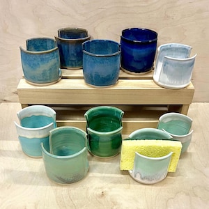 May include: A collection of ceramic sponge holders in various shades of blue, green, and white, arranged on a wooden tiered stand. Each holder features a U-shaped cutout on one side, and one holder contains a rectangular yellow kitchen sponge.