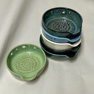 May include: A collection of six ceramic graters in various shades of blue, green, and white. Each grater features a textured grating surface and a small spout. The top grater is a deep teal, and the bottom grater is a light green.