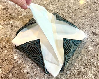 May include: A square ceramic napkin holder with a dark teal, glossy glaze and a textured, leaf-like pattern on the exterior. The holder contains a stack of folded white paper napkins, with one napkin being pulled upward from the center.
