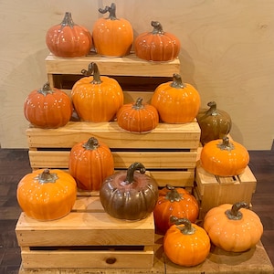 May include: A collection of fourteen ceramic pumpkins in various shades of orange and brown, each featuring a glossy finish and a dark, curved stem. The pumpkins are arranged on stacked wooden crates of varying heights.