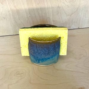 May include: A ceramic sponge holder with a glossy glaze that transitions from deep navy blue at the top to a lighter turquoise at the base. A rectangular yellow kitchen sponge with a dark green scouring layer is inserted into the holder.