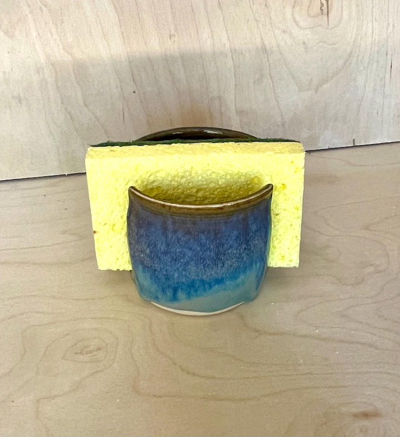 May include: A ceramic sponge holder with a glossy glaze that transitions from deep navy blue at the top to a lighter turquoise at the base. A rectangular yellow kitchen sponge with a dark green scouring layer is inserted into the holder.