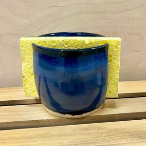 May include: A deep blue ceramic sponge holder with a glossy finish, featuring a curved, open-top design. A rectangular yellow porous sponge is inserted into the holder, extending beyond the top and sides of the blue ceramic container.