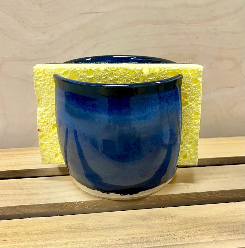 May include: A deep blue ceramic sponge holder with a glossy finish, featuring a curved, open-top design. A rectangular yellow porous sponge is inserted into the holder, extending beyond the top and sides of the blue ceramic container.
