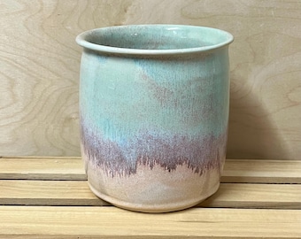 May include: A cylindrical ceramic mug featuring a smooth, rounded rim. The exterior glaze transitions from a soft, pale mint green at the top to a textured, dusty mauve midsection, fading into a warm, creamy beige at the base.