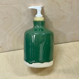 Pu&ograve; includere: Un dispenser per sapone in ceramica con una sezione superiore smaltata in verde scuro lucido e una base opaca bianca non smaltata. Il contenitore presenta un corpo cilindrico con un collo stretto e un meccanismo a pompa in plastica bianca.