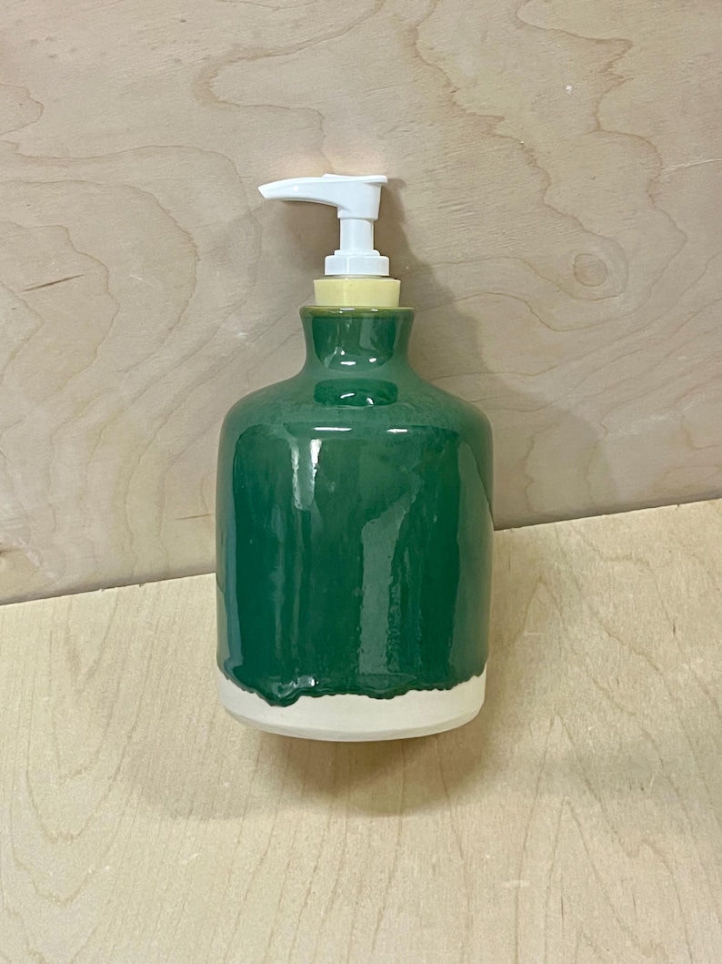 Pu&ograve; includere: Un dispenser per sapone in ceramica con una sezione superiore smaltata in verde scuro lucido e una base opaca bianca non smaltata. Il contenitore presenta un corpo cilindrico con un collo stretto e un meccanismo a pompa in plastica bianca.