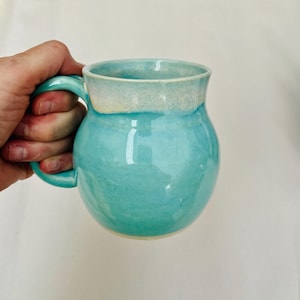 May include: A handmade ceramic mug in a vibrant turquoise glaze. The mug features a rounded body, a sturdy handle, and a contrasting off-white rim. The mug is held in a hand, showcasing its size and shape. This unique mug is perfect for coffee or tea.