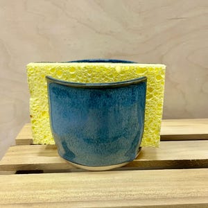 May include: A blue ceramic sponge holder with a glossy, speckled glaze finish. The container features a curved front edge and holds a rectangular yellow kitchen sponge. The ceramic piece has a smooth, rounded base and a deep blue color.
