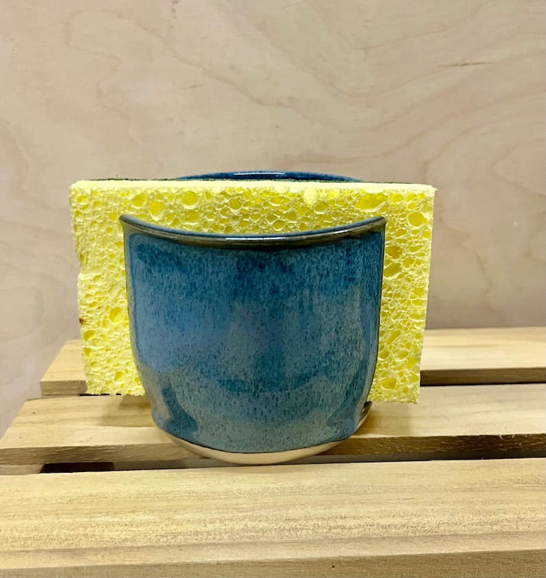 May include: A blue ceramic sponge holder with a glossy, speckled glaze finish. The container features a curved front edge and holds a rectangular yellow kitchen sponge. The ceramic piece has a smooth, rounded base and a deep blue color.