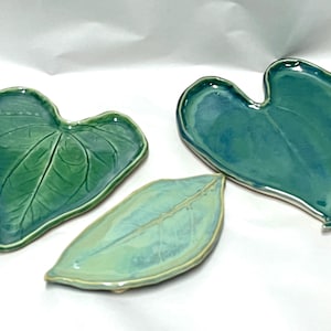 May include: Three ceramic dishes shaped like leaves. Two dishes are heart-shaped with deep green and teal glossy glazes, featuring embossed vein details. The third dish is an elongated oval shape in a lighter, pale mint green glaze with visible veins.