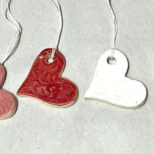 May include: Close-up of three ceramic heart ornaments. One is red with a floral design, another is white, and a third is pink. Each heart has a hole at the top for hanging with a white string. The ornaments are on a light gray surface.