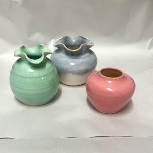 May include: Three ceramic vases in shades of green, blue, and pink. The green vase has a ruffled rim and horizontal ridges. The blue vase has a ruffled rim and a gradient of blue to white. The pink vase is smooth and round.
