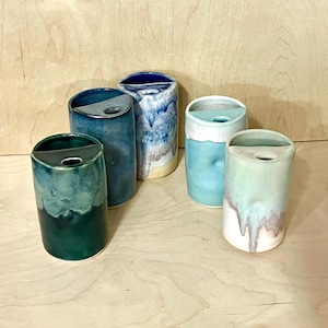 May include: Five cylindrical ceramic vases with a semi-circular top opening and a small circular hole. The vases feature various glaze finishes in shades of deep forest green, navy blue, mottled blue and white, light turquoise, and pale mint green.