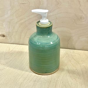 Pu&ograve; includere: Un dispenser per sapone in ceramica con una finitura lucida nei toni del verde acqua e verde. Il corpo cilindrico presenta sottili scanalature orizzontali e una base liscia non smaltata. Un erogatore a pompa in plastica bianca &egrave; fissato al collo.