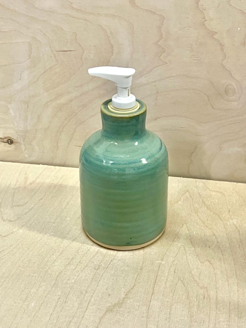 Pu&ograve; includere: Un dispenser per sapone in ceramica con una finitura lucida nei toni del verde acqua e verde. Il corpo cilindrico presenta sottili scanalature orizzontali e una base liscia non smaltata. Un erogatore a pompa in plastica bianca &egrave; fissato al collo.