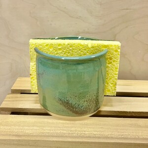May include: A ceramic sponge holder with a glossy finish in shades of teal, green, and brown. The container features a curved, open-top design and holds a rectangular yellow kitchen sponge with a green scouring pad on the back side.
