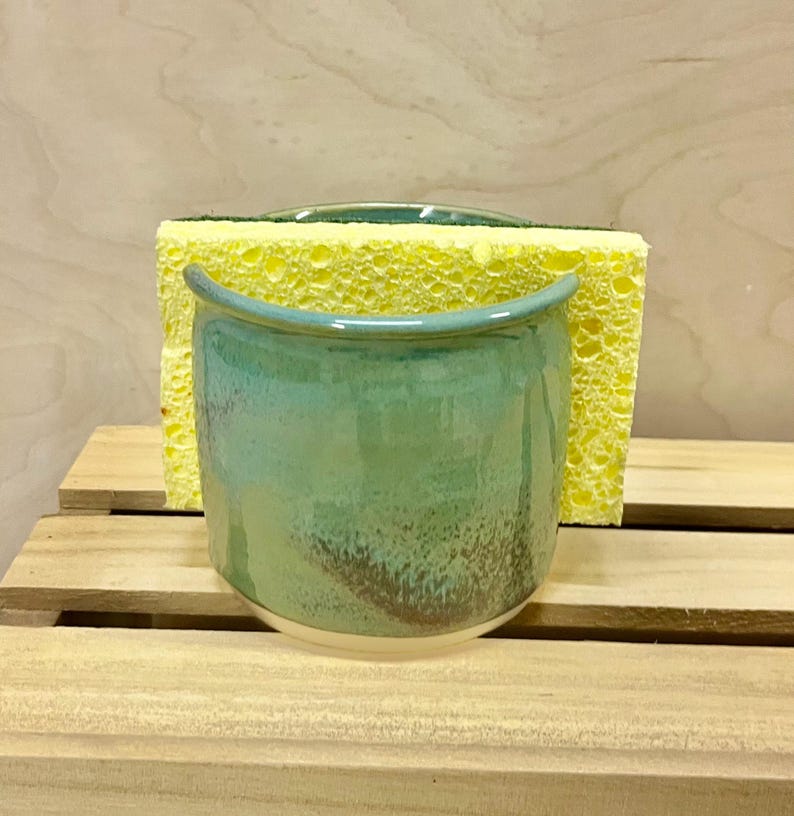 May include: A ceramic sponge holder with a glossy finish in shades of teal, green, and brown. The container features a curved, open-top design and holds a rectangular yellow kitchen sponge with a green scouring pad on the back side.
