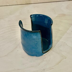 May include: A ceramic sponge holder with a glossy blue glaze and a dark rim. The vessel features a wide, U-shaped cutout on one side, allowing for easy access to the interior. The exterior surface shows subtle variations in the blue color tones.