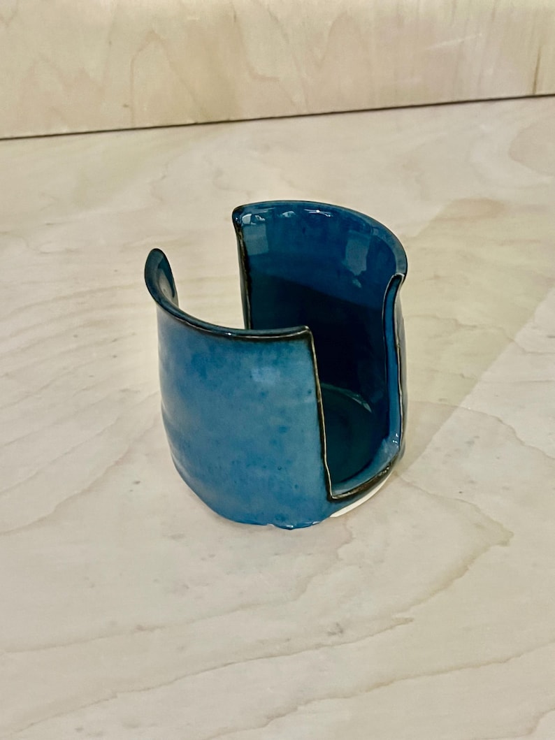 May include: A ceramic sponge holder with a glossy blue glaze and a dark rim. The vessel features a wide, U-shaped cutout on one side, allowing for easy access to the interior. The exterior surface shows subtle variations in the blue color tones.
