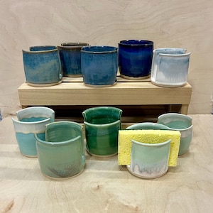 May include: A collection of ten ceramic sponge holders in various shades of blue, green, and white. Each holder features a U-shaped cutout on the front rim. One holder contains a rectangular yellow sponge. The items are arranged on two levels.