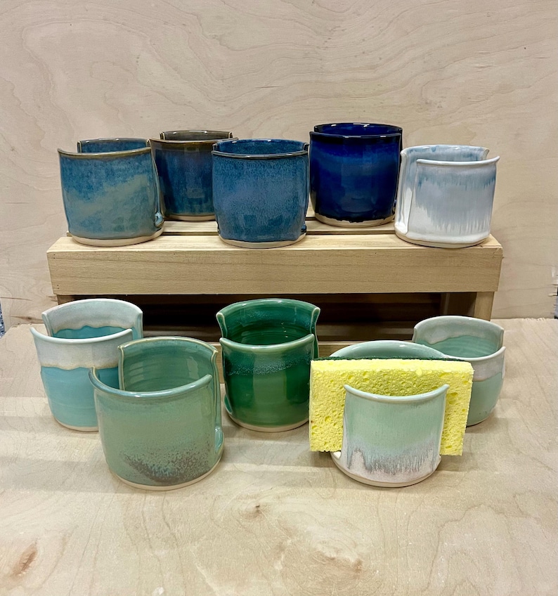 May include: A collection of ten ceramic sponge holders in various shades of blue, green, and white. Each holder features a U-shaped cutout on the front rim. One holder contains a rectangular yellow sponge. The items are arranged on two levels.