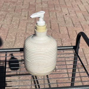 Pu&ograve; includere: Un dispenser di sapone in ceramica color crema con un corpo arrotondato e scanalato e un meccanismo a pompa in plastica bianca. Il dispenser presenta una superficie strutturata con scanalature orizzontali e un collo cilindrico liscio.