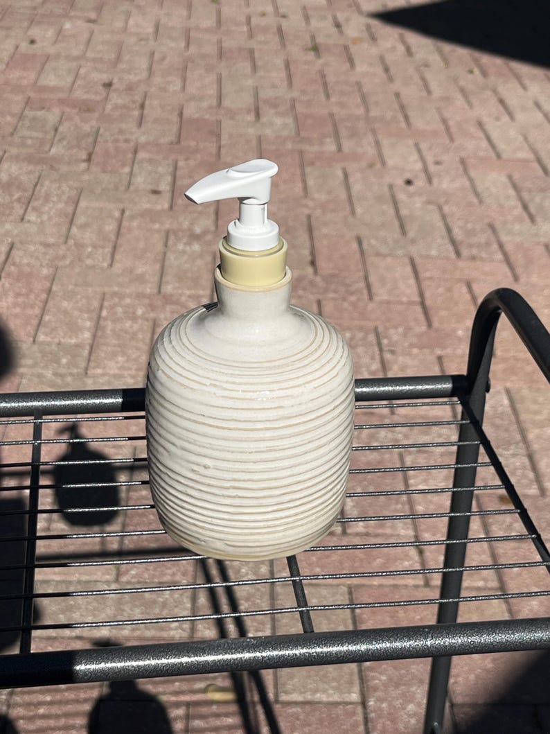 Pu&ograve; includere: Un dispenser di sapone in ceramica color crema con un corpo arrotondato e scanalato e un meccanismo a pompa in plastica bianca. Il dispenser presenta una superficie strutturata con scanalature orizzontali e un collo cilindrico liscio.