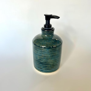 Pu&ograve; includere: Un dispenser di sapone cilindrico in ceramica caratterizzato da uno smalto reattivo blu e verde acqua profondo con una texture a coste orizzontali. Il contenitore &egrave; dotato di una pompa in plastica nera che contrasta con la finitura lucida.