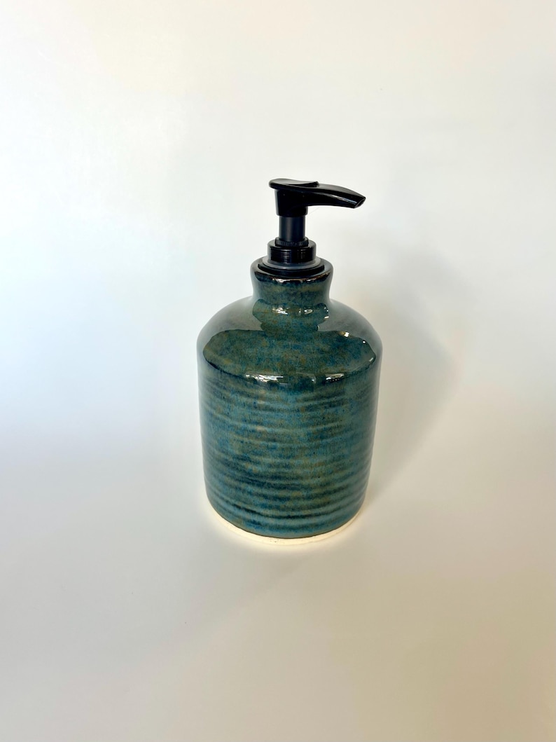 Pu&ograve; includere: Un dispenser di sapone cilindrico in ceramica caratterizzato da uno smalto reattivo blu e verde acqua profondo con una texture a coste orizzontali. Il contenitore &egrave; dotato di una pompa in plastica nera che contrasta con la finitura lucida.