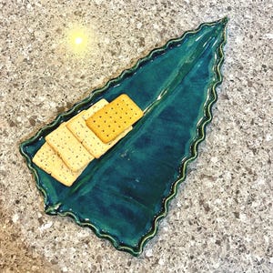 May include: A glossy, deep teal ceramic serving tray shaped like a long, pointed leaf with a ruffled, wavy edge. The tray features a central vein detail and holds a small stack of four rectangular, light brown crackers on one side.