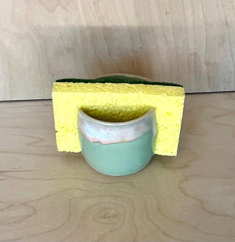 May include: A small, round, light green ceramic holder with a smooth glaze and a pale pink rim. A rectangular yellow sponge with a dark green scouring layer is inserted into the holder, resting against the curved front of the ceramic piece.
