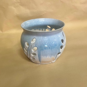 May include: A round ceramic planter with a glossy finish featuring a gradient of light blue and white. The vessel has a wide opening and multiple oval-shaped cutouts along the sides, allowing for ventilation and visibility through the structure.