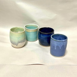 May include: Four ceramic cups with rounded bodies and smooth rims. One cup features a pale green and white gradient, another is a solid light turquoise, the third is a deep navy blue, and the fourth is a periwinkle blue with a white drip glaze.